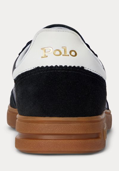 Zwarte suède sneakerhiel met witte leren bovenkant en gouden "Polo"-logo boven bruine gestructureerde rubberen zool.