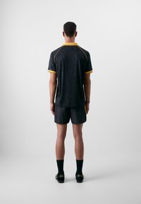 T-shirt de sport noir avec des accents jaunes et des motifs subtils, associé à un short et des chaussettes noirs, vu de dos.