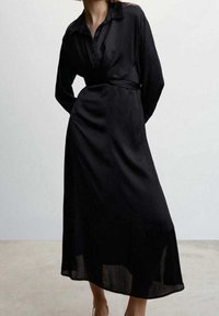 Robe noire à manches longues avec un col à revers, une ceinture à la taille et un ourlet fluide. Fabriquée en tissu léger avec une texture légèrement transparente.