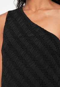 Melns tops ar teksturētu audumu, kas rotāts ar diagonālām svītrām un maziem izceltiem rakstiem. Asimetrisks kakla izgriezums, gluda virsma un viegla drapēšana.