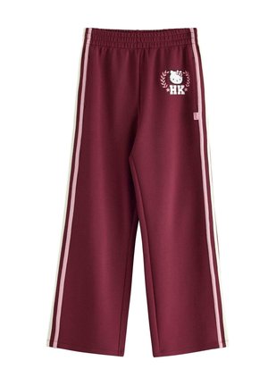 Młoda kobieta o długich blond włosach, ubrana w białą koszulkę Hello Kitty oraz bordowe spodnie z logo Hello Kitty, stoi z rękami w kieszeniach.
