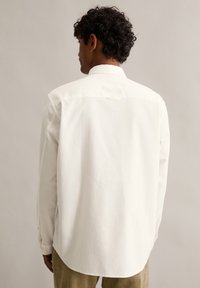 Biała koszula z długim rękawem, z klasycznym kołnierzem, karczkiem na plecach i zaokrąglonym dołem. Materiał wydaje się gładki z delikatnym połyskiem.