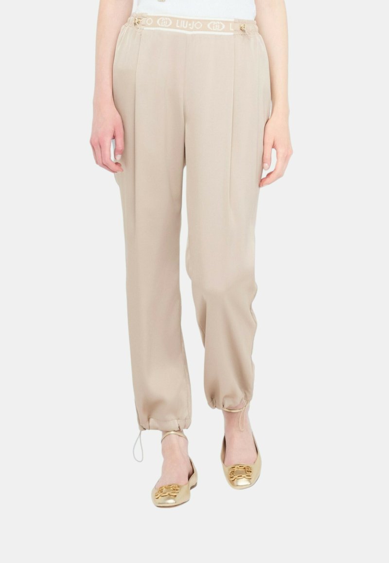 Pantaloni leggeri beige con vita elasticizzata e stringa. Presentano gambe tapered con polsini elasticizzati. Indossati con ballerine dorate.