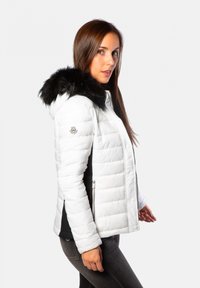 Doudoune blanche avec une capuche en fausse fourrure noire, présentant des coutures horizontales, une poche latérale zippée et une coupe ajustée.
