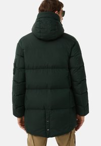 Mørkegrøn puffjakke med hætte, der har et quiltet design, synlige sømme og et logo på ryggen. Glat, isoleret tekstur.