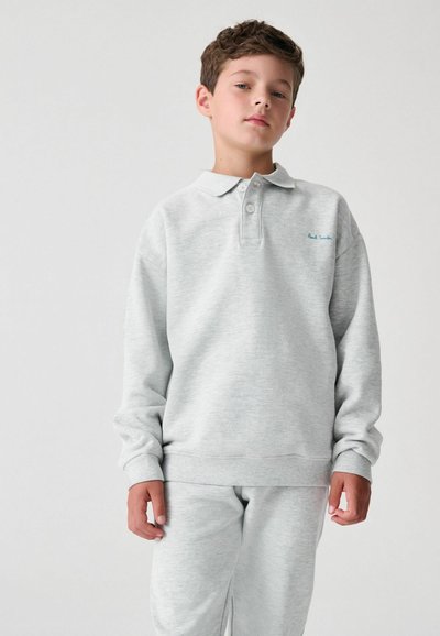 Šviesiai pilkas džemperis su polo apykakle, turintis tris mygtukus ir raštuotas rankogalius. Dėvimas su atitinkančiomis pilkomis treniruotėmis. Mėlynu siuvinėjimu užrašas.