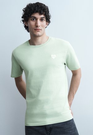 Jeune homme aux cheveux bouclés et foncés, portant une chemise à manches courtes vert clair avec un petit emblème en forme de cœur, debout devant un fond clair uni.