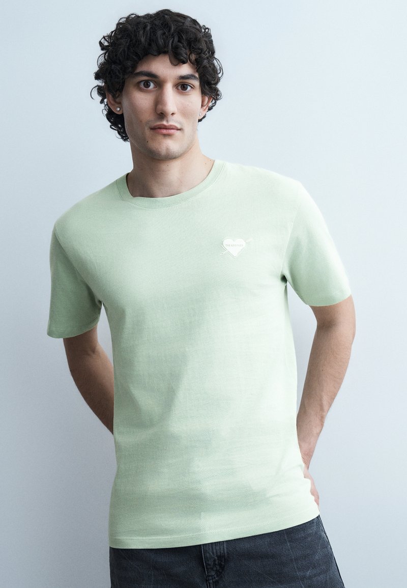 Νεαρός άνδρας με σγουρά σκούρα μαλλιά, φορώντας ανοιχτοπράσινο κοντομάνικο πουκάμισο με μικρό έμβλημα καρδιάς, στέκεται μπροστά σε απλό ανοιχτό φόντο.