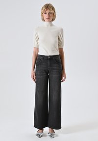 Een vrouw staat in een witte korte mouwen turtlenecktop, hoge taille zwarte wijde jeans en zilveren puntige schoenen met strikken.