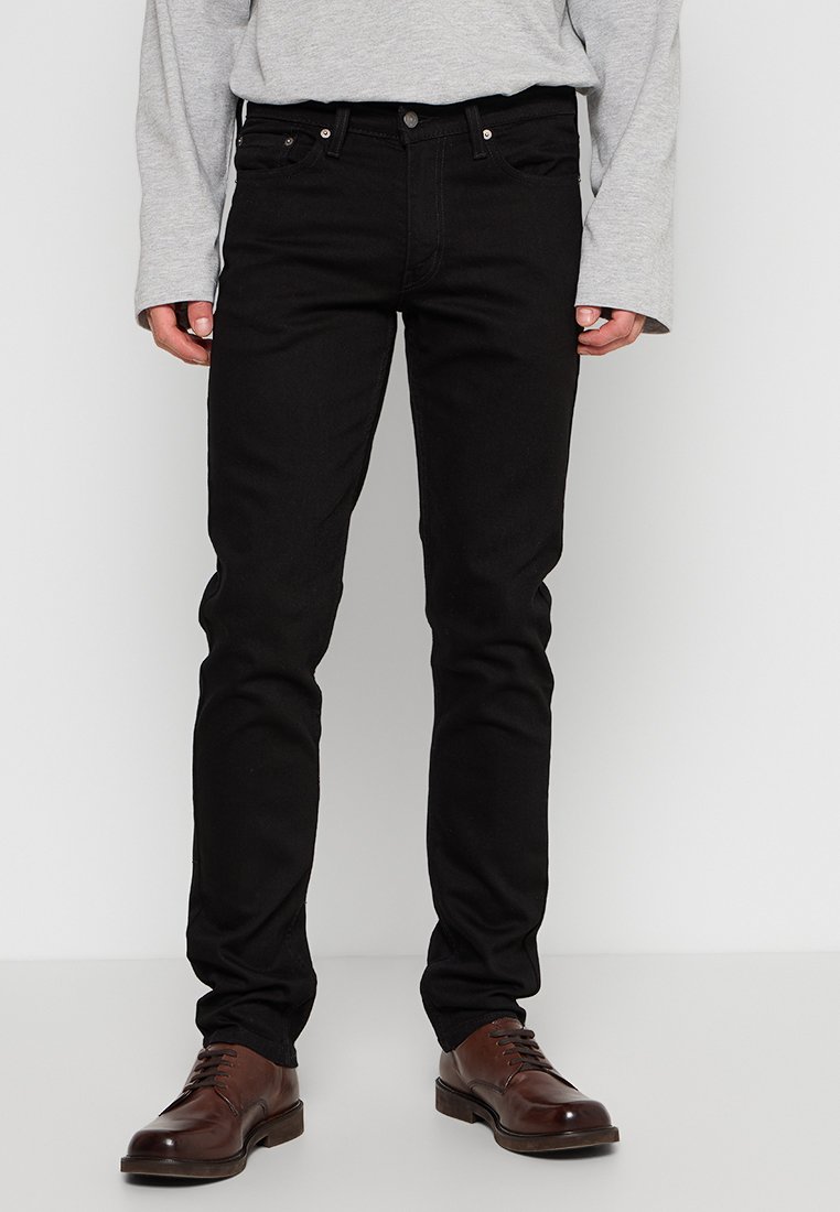 Jeans de mezclilla negra en corte ajustado con cinco bolsillos, con herrajes plateados y una textura suave. Combinados con una camiseta de manga larga gris y zapatos marrones.