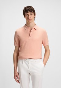 Jovem vestido com uma camisola polo de manga curta rosa clara e calças brancas, de pé com uma mão no bolso, num fundo simples.