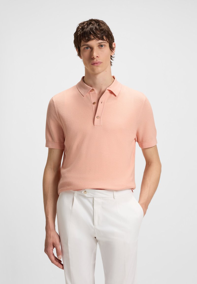 Jovem vestido com uma camisola polo de manga curta rosa clara e calças brancas, de pé com uma mão no bolso, num fundo simples.