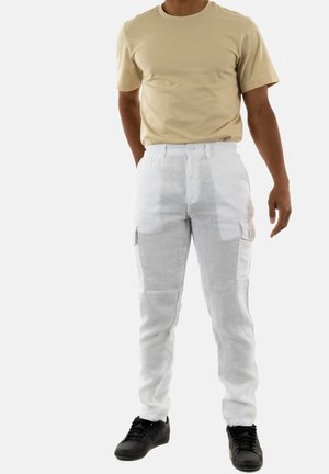 Beige Baumwoll-T-Shirt kombiniert mit weißen Leinen-Cargos, die seitliche Taschen und eine leicht strukturierte Oberfläche aufweisen. Schwarze Sneakers runden den Look ab.