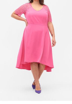 Vrouw in een felroze knielange jurk met kanten mouwen en paarse hoge hakken, staand met een hand op haar heup.