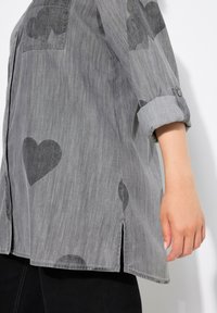 Camicia grigia con maniche arrotolate, decorata con motivi di cuori neri e nuvole; le fessure laterali migliorano il flusso d'aria e il movimento.