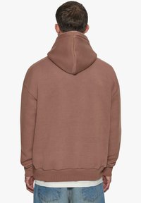 Brun oversized hoodie med snoretræk i hætten, ribbede manchetter og kant. Blød tekstur med en afslappet pasform, båret over en hvid T-shirt.