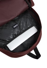 Interno di uno zaino marrone con fodera a scacchi nera, compartimento imbottito per un laptop e etichetta con il logo Vans. Chiusura con zip.