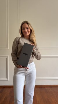 Mlada ženska v bež puloverju in belih hlačah drži sivo škatlo z oznako "BRONS" v prostoru s smetanastimi stenami in lesenim podom.