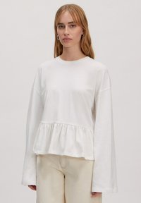 Witte langslipshirt met een geplooide peplumzoom, gemaakt van zachte stof. Heeft een ronde halslijn en een relaxte pasvorm. Gecombineerd met lichtbeige broek.