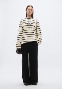 Oversized sweatshirt met crème- en marineblauwe strepen en logo, gecombineerd met zwarte broek met wijde pijpen. Model houdt een kleine zwarte tas vast.