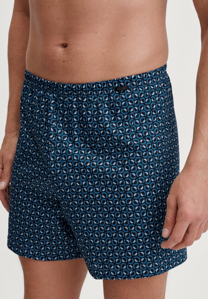 Blauwe zwemshorts met een geometrisch patroon, een elastische tailleband en een gladde stoftextuur. Merktag zichtbaar op de tailleband.