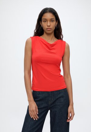Femme aux longs cheveux foncés portant un haut rouge sans manches et un jean bleu foncé, debout devant un fond blanc uni.
