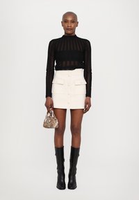 Schwarzes, transparentes Langarmoberteil, weißer Minirock mit Knöpfen vorne und Taschen, kniehohe schwarze Stiefel, kleine gemusterte Handtasche.