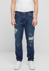 Jeans de mezclilla azul con corte recto, con desgastes visibles y parches, complementados con costuras naranjas y cierre de botón.