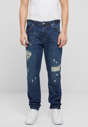 Jeans en denim bleu avec une coupe droite, présentant des déchirures et des réparations visibles, agrémentés de surpiqûres orange et d'une fermeture à bouton.