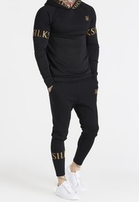 Chándal negro deportivo con una parte superior con capucha de manga larga y pantalones ajustados, con detalles dorados en las mangas y las piernas, combinado con zapatillas blancas.