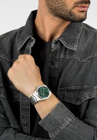Orologio da polso in argento con quadrante verde, cinturino in acciaio inossidabile e componenti in tono argento. Indossato su una mano, parzialmente coperto da una giacca di jeans.