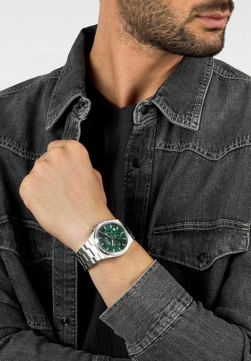 Orologio da polso in argento con quadrante verde, cinturino in acciaio inossidabile e componenti in tono argento. Indossato su una mano, parzialmente coperto da una giacca di jeans.