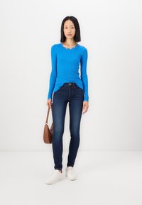 Blauwe lange mouwen top met een scalloped halslijn, gecombineerd met donkerblauwe skinny jeans en witte sneakers. Bruine schoudertas als accessoire.