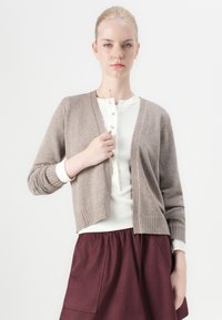 Bruin gebreide cardigan met V-hals, geribbelde manchetten en zoom, gelaagd over een witte overhemd met knopen, gecombineerd met een kastanjebruine rok.