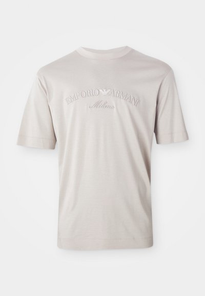 Camiseta de manga corta color beige claro, hecha de una tela suave, con el logo bordado "Emporio Armani Milano" en un tono ligeramente más oscuro.