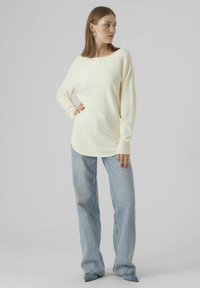 Cremefarbener Strickpullover mit weitem Rundhalsausschnitt, langen Ärmeln und lockerer Passform, kombiniert mit hellblauen Jeans und spitz zulaufenden Schuhen.