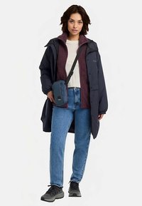 Donkerblauwe lange jas over een mahoniekleurige fleece, beige overhemd, lichtblauwe spijkerbroek en zwarte sneakers, met een kleine blauwe crossbodytas.