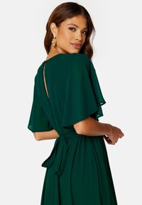 Robe verte fluide avec des manches courtes et flottantes et un détail en forme de trou de serrure dans le dos. Présente une taille nouée et un tissu lisse et léger.