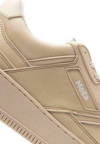 Bežs ikdienas sporta apavi ar teksturētu zoli, izšūtām ādas paneļiem un baltu "MOEa" logotipu sānos.