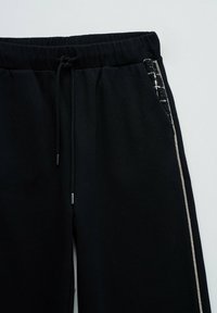 Pantalons de survêtement noirs avec taille élastique, cordon de serrage et une bande décorative en strass argentés le long de la couture latérale.