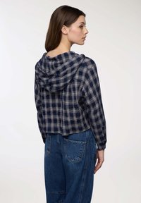 Giovane donna in camicia a scacchi blu navy e grigia con cappuccio e jeans di denim blu, posata di lato con la schiena per lo più visibile.
