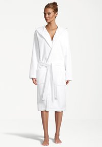 Vrouw die blootsvoets staat in een witte badjas met capuchon, vastgebonden op de taille met twee voorkeurszakken, tegen een effen achtergrond.