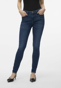 Jeans ajustados de color azul oscuro con una cintura de altura media, diseño de cinco bolsillos y un ligero desvanecimiento. Acompañados de zapatos de tacón negros con punta afilada.