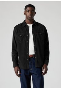 Camisa preta de veludo cotelê com fecho de botões, dois bolsos no peito e gola de bico. Combinada com jeans de ganga azul, usada com cinto castanho.