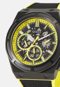 Reloj negro con una correa de silicona amarilla texturizada, que presenta un dial esqueletizado, tres sub-diales y acentos en plata y amarillo contrastantes.