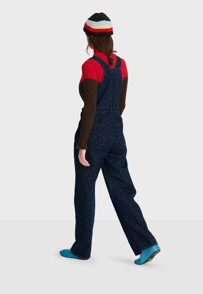 Femme qui s'éloigne portant une salopette bleu foncé à motifs, un pull côtelé rouge et marron, un bonnet rayé multicolore et des chaussures bleues.