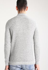 Pull gris tricoté avec une finition texturée, col rond et manches longues. Comprend un ourlet et des poignets côtelés, porté avec un jean bleu clair.