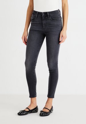Dark gray skinny jeans with a high waist, featuring front pockets and a smooth texture. Paired with black studded flats.