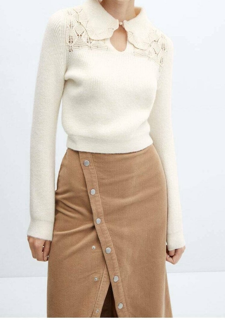 Femme portant un pull crème en maille avec un col en dentelle et une jupe en velours côtelé beige à boutons, debout devant un fond uni.