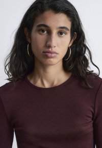 Burgunderfarbenes Langarmoberteil mit Rundhalsausschnitt aus strukturiertem Stoff, getragen von einer Person mit dunklem, welligem Haar und goldenen Creolen.